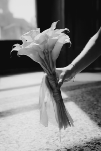 Eine Hand hält einen Calla-Strauß, elegant mit einer Schleife umwickelt, beleuchtet von sanftem Tageslicht, das durch ein nahes Fenster fällt. Der leicht verschwommene Hintergrund betont die Eleganz des Hochzeitsarrangements. Schwarzweißbild.