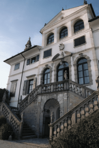 Eine große, elegante Villa, perfekt für eine Traumhochzeit, mit einer großen Steintreppe zum Eingang. Die Fassade zeichnet sich durch hohe Bogenfenster, weiße Wände und kunstvolle Details aus. Skulpturen säumen die Treppe vor einem klaren blauen Himmel. Ein atemberaubender Ort für Ihren besonderen Tag.
