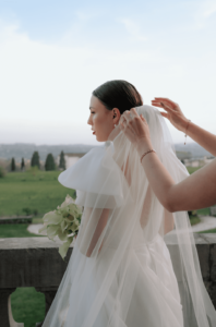 Eine Braut in weißem Kleid und Schleier, ein Bild eleganter Hochzeitsglückseligkeit, steht auf einem Balkon mit Blick auf eine grüne Wiese. Jemandes Hände richten sanft ihren Schleier, während sie einen Blumenstrauß hält. Der Himmel ist klar und hell und spiegelt den freudigen Geist von Heiraten wider.