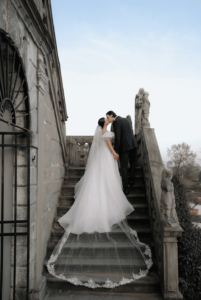 Braut und Bräutigam küssen sich auf einer alten Steintreppe und halten damit einen perfekten Moment ihrer Hochzeit fest. Die Braut trägt ein fließendes weißes Kleid mit langer Schleppe, während der Bräutigam einen dunklen Anzug trägt. Sie stehen auf verzierten Stufen, die mit Steinfiguren gesäumt sind, unter einem klaren blauen Himmel.
