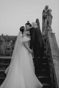 Braut und Bräutigam küssen sich auf einer Steintreppe, umgeben von Statuen, und verkörpern damit eine klassische Hochzeit. Die Braut trägt ein schulterfreies Kleid mit langem Schleier, während der Bräutigam einen dunklen Anzug mit weißen Blumen in der Tasche trägt. Die Szene ist zeitlos romantisch, perfekt für Heiratserinnerungen.