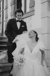 Ein Schwarzweißfoto eines Paares an seinem Hochzeitstag fängt die Essenz von Liebe und Eleganz ein. Die lächelnde Braut hält einen Blumenstrauß und trägt ein wallendes Kleid. Der Bräutigam im Smoking steht hinter ihr auf Steinstufen vor einem Gebäude mit großen Fenstern.