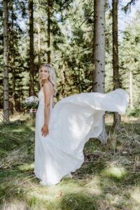 Eine Frau in einem fließenden weißen Brautkleid steht auf einer Waldlichtung und hält einen Blumenstrauß in der Hand. Während das Sonnenlicht durch die Bäume fällt und eine heitere und magische Atmosphäre schafft, stellt sie sich ihre perfekte Hochzeit inmitten der Natur vor.