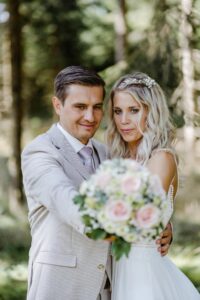 Braut und Bräutigam posieren während ihrer Hochzeit im Freien und halten einen Strauß rosa und weißer Blumen in den Händen. Der Bräutigam trägt einen hellgrauen Anzug, während die Braut ein weißes Kleid trägt und ihr Haar elegant geschmückt ist. Sie sind von üppigem Grün umgeben.