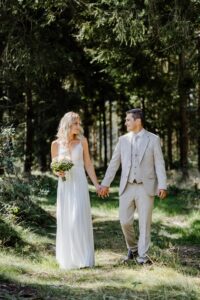 Braut und Bräutigam gehen Hand in Hand durch einen sonnenbeschienenen Wald und verkörpern damit die Essenz einer perfekten Hochzeit. Die Braut trägt ein weißes Kleid und hält einen Blumenstrauß, während der Bräutigam einen hellgrauen Anzug trägt. Beide schauen sich inmitten des üppigen Grüns an.