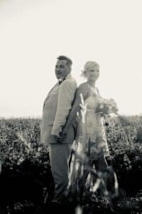 Inmitten ihrer sorgfältigen Hochzeitsplanung stehen Braut und Bräutigam Rücken an Rücken auf einem sonnenbeschienenen Feld. Der Bräutigam trägt einen hellen Anzug, während die Braut in ihrem Spitzenkleid mit einem Blumenstrauß in der Hand von der strahlenden Sonne umrahmt wird – ein perfekter Moment, den sie auf ihrer Reise nach Heiraten genießen können.