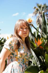 Eine junge Frau mit langen Haaren steht vor tropischen Pflanzen mit großen, orangefarbenen Blüten und trägt ein geblümtes Kleid, während sie sich ihre Traumhochzeit vorstellt. Im Hintergrund sind ein klarer blauer Himmel und eine große Palme zu sehen, die die ruhige Kulisse ihrer Hochzeitsplanung perfekt ergänzen.