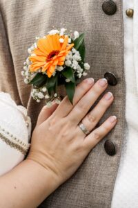 Nahaufnahme einer Person, die einen Ehering an der linken Hand trägt und ihn einer anderen Person auf die Brust legt. Die Person im Hintergrund trägt ein beiges Jackett und eine orangefarbene Ansteckblume mit Blumenmotiv, die Liebe und Heirat symbolisiert. Der Fokus liegt auf den gefalteten Händen und dem Blumendetail.