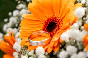 Eine Nahaufnahme eines silbernen Eherings ruht auf leuchtend orangefarbenen Gerbera-Gänseblümchen, umgeben von zarten weißen Schleierkrautblüten. Der unscharfe Hintergrund hebt die hellen Farben und komplizierten Details hervor, perfekt für jede Hochzeitsplanung.