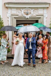 Eine fröhliche Braut und ein fröhlicher Bräutigam gehen Hand in Hand unter einem Regenschirm, umgeben von lächelnden Gästen, die ihre eigenen Regenschirme halten. Die Hochzeitskulisse zeigt einen eleganten Gebäudeeingang mit kunstvollen Details. Die Szene ist trotz des regnerischen Wetters lebhaft und fröhlich, ein Beweis für eine durchdachte Hochzeitsplanung.