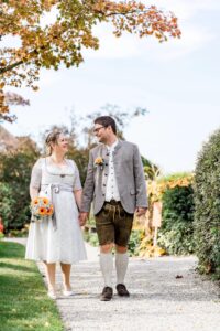 Ein Paar in traditioneller Kleidung geht Hand in Hand einen Kiesweg entlang, umgeben von Grün und Herbstlaub. Die Frau trägt einen Strauß Sonnenblumen und lächelt den Mann an. Der Himmel ist klar und sonnig. Es ist ein perfekter Tag für eine unvergessliche Hochzeit.