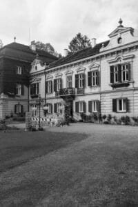 Ein Schwarzweißbild zeigt ein großes, altes europäisches Herrenhaus, perfekt für die Hochzeitsplanung. Die kunstvollen architektonischen Details und der dekorative Balkon strahlen zeitlose Eleganz aus. Ein Weg führt zum Eingang und lässt auf unvergessliche Hochzeitsmomente schließen, wobei der Vorgarten teilweise sichtbar ist.