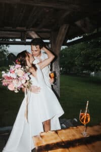Ein Paar in Hochzeitskleidung teilt einen romantischen Moment unter einer Holzkonstruktion und verkörpert damit den Geist der Hochzeit. Die Braut hält einen bunten Blumenstrauß in der Hand, während der Bräutigam sein Getränk hält. Mit üppigem Grün und Bäumen im Hintergrund ist dies eine bezaubernde Szene, perfekt für jeden, der sich intensiv mit der Hochzeitsplanung beschäftigt.