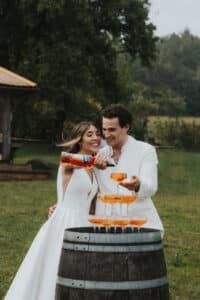 Ein Paar in eleganter weißer Kleidung gießt orangefarbene Getränke in einen Turm aus Champagnergläsern, die draußen auf einem Fass stehen. Lächelnd feiern sie ihre Hochzeit inmitten einer üppigen grünen Umgebung mit Bäumen im Hintergrund. Die Freude ist in jedem Moment dieses bezaubernden Tages spürbar.