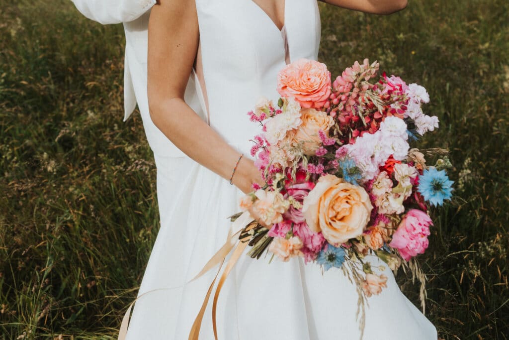 Eine Braut in einem weißen Kleid hält einen leuchtend bunten Blumenstrauß, darunter rosa, orange und blaue Blüten. Sie steht auf einer Wiese, ist teilweise sichtbar, hat den Rücken leicht abgewandt und verkörpert die Magie von Hochzeitsträumen.