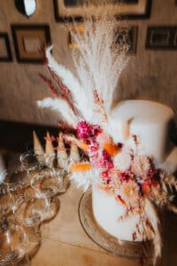 Eine weiße Torte, geschmückt mit getrockneten Blumen und Federn, steht auf einem Holztisch neben einer Reihe leerer Champagnergläser – ein perfektes Herzstück jeder Hochzeitsplanung. Der Hintergrund besteht aus einer strukturierten Wand mit gerahmten Bildern, die der Hochzeitsfeier eine elegante Note verleihen.