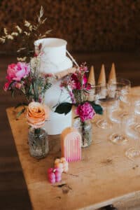 Ein rustikaler Tisch, geschmückt mit rosa und orangefarbenen Blumen in Vasen, einem weißen Krug, einzigartig geformten Kerzen, klaren Weingläsern und Holzkegeln, schafft die perfekte Kulisse für eine warme, elegante Hochzeitsatmosphäre.