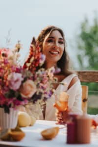 Eine Frau mit langen Haaren sitzt lächelnd an einem Tisch im Freien und plant möglicherweise ihre Hochzeit. In einer Hand hält sie ein Getränk, daneben steht ein bunter Blumenstrauß und Früchte wie Zitronen und Orangen. Im Hintergrund ist eine verschwommene grüne Landschaft zu sehen.