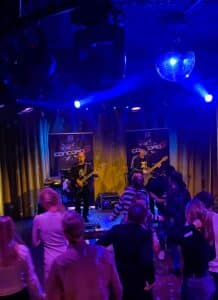 Eine kleine Band tritt auf einer schwach beleuchteten Bühne in einem Veranstaltungsort namens „Concord“ auf und verleiht einer Hochzeitsfeier Magie. Zwei Musiker spielen Gitarre unter violettem und blauem Bühnenlicht, während die Gäste energisch tanzen. Eine Discokugel hängt von der Decke und verstärkt die festliche Atmosphäre dieses besonderen Tages.