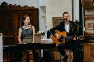 Eine Frau und ein Mann musizieren drinnen; die Frau spielt Keyboard und singt, während der Mann eine Akustikgitarre klimpert. Ihre harmonischen Melodien erfüllen einen Raum mit Holzmöbeln und Steinwänden und erzeugen die warme Atmosphäre, die perfekt für eine intime Hochzeitsfeier ist.