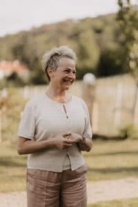 Eine lächelnde Frau mit kurzen Haaren steht im Freien und trägt ein helles Hemd und eine helle Hose. Sie befindet sich in einer grünen, natürlichen Umgebung mit verschwommenen Bäumen und Gras im Hintergrund und stellt sich vielleicht ihre bevorstehende Hochzeit inmitten der bezaubernden Umarmung der Natur vor.