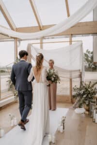 Braut und Bräutigam gehen im Zauber einer Hochzeit durch einen Innengang auf einen Geistlichen zu. Die Braut trägt ein langes weißes Kleid mit offenem Rücken und der Bräutigam einen blauen Anzug. Der elegant dekorierte Raum ist mit transparenten Stoffen, Pflanzen und Kerzen ausgestattet, während natürliches Licht durch große Fenster strömt.