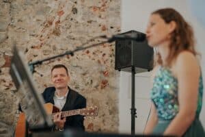 Ein Mann spielt Akustikgitarre und lächelt, während er vor einer Steinmauer sitzt und so die perfekte Atmosphäre für eine Hochzeit schafft. In der Nähe steht eine Frau in einem glitzernden Kleid neben einem Mikrofon, leicht verschwommen – und fängt die Essenz einer intimen Hochzeitsplanung bei einer Veranstaltung in geschlossenen Räumen ein.