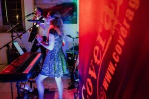 In einem warm beleuchteten Raum spielt eine Band und erweckt Melodien zum Leben. Eine Frau singt und spielt Keyboard, während ein Mann auf seiner Gitarre klimpert und ins Mikrofon harmonisiert. Ein großes Banner mit dem Wort „Cover“ weist auf ihre besondere Rolle bei Ihrer Hochzeitsplanung hin.