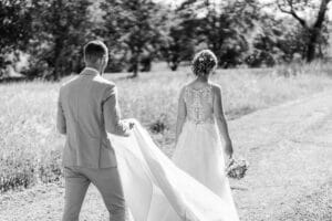 Ein Schwarzweißfoto zeigt ein Paar auf seiner Hochzeitsreise einen Feldweg entlang. Die Braut in einem langen Spitzenkleid hält einen Blumenstrauß und wendet sich leicht ab, während der Bräutigam in einem hellen Anzug sanft ihre Schleppe hält. Bäume und Gras umgeben liebevoll ihre romantische Kulisse.