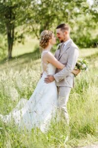 Braut und Bräutigam stehen auf einer Wiese, umarmen sich und lächeln sich an ihrem Hochzeitstag an. Die Braut trägt ein weißes Kleid mit Spitzendetails, während der Bräutigam einen hellen Anzug trägt und einen Blumenstrauß hält. Im Hintergrund sind Bäume unter einem sonnigen Himmel zu sehen.