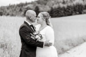Braut und Bräutigam stehen eng beieinander auf einem Feldweg und blicken einander in die Augen. Dies verkörpert die Essenz einer Hochzeit. Die Braut hält einen Blumenstrauß. Im Hintergrund sind Felder und Bäume zu sehen, die in diesem Schwarzweißbild eine ruhige, romantische Atmosphäre schaffen.