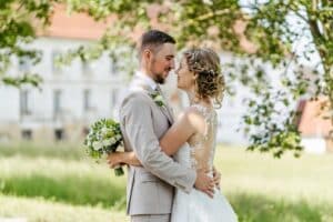 Ein Paar im Hochzeitskleid steht eng umschlungen im Freien und verkörpert seinen Hochzeitstraum. Der Bräutigam trägt einen hellgrauen Anzug und die Braut ein weißes Spitzenkleid und hält einen Blumenstrauß in der Hand. Sie blicken einander in die Augen, während Bäume und ein Gebäude ihren Moment der Liebe sanft einrahmen.