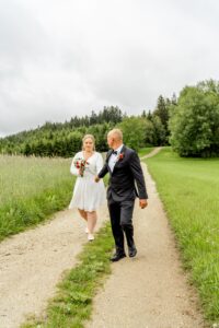 Ein Paar spaziert auf einem Kiesweg, umgeben von grünem Gras und Bäumen, und fängt die Essenz ihres Hochzeitstages ein. Die Frau im weißen Kleid hält einen Strauß roter und weißer Blumen in der Hand. Neben ihr geht der Mann im dunklen Anzug unter einem bedeckten Himmel und besiegelt ihre Liebe in perfekter Harmonie.