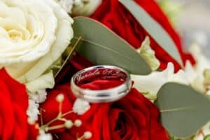 Ein silberner Ring mit der Gravur „Daniela 02-07-2022“ ruht auf einem Strauß aus roten Rosen, weißen Rosen und grünen Blättern – eine perfekte Momentaufnahme sorgfältiger Hochzeitsplanung.