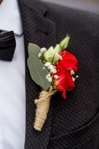 Eine Nahaufnahme einer Person, die einen dunkelkarierten Anzug mit weißem Hemd und schwarzer Fliege trägt, perfekt für eine elegante Hochzeit. Eine Ansteckblume mit roten und weißen Rosen, Schleierkraut und grünen Blättern ist mit Schnur umwickelt und am Revers befestigt.