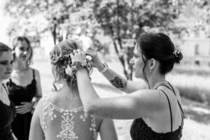 Eine Frau steckt einer Braut, die im Freien ein Spitzenkleid trägt, ein Blumenaccessoire ins Haar und verkörpert damit die Essenz einer perfekten Hochzeit. Zwei weitere Frauen in eleganter Kleidung stehen daneben. Die Kulisse ist ein sonniger Tag mit Bäumen und Gras im Hintergrund, ideal für solche Hochzeitsfeierlichkeiten.