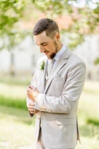 Ein Mann in einem hellbeigen Anzug steht während einer Hochzeit im Freien auf einer sonnenbeschienenen Wiese. Er rückt seinen Ärmel zurecht, trägt ein weißes Hemd und eine graue Krawatte und hat eine Ansteckblume am Revers. Der Hintergrund ist sanft verschwommen und zeigt Bäume und ein Gebäude, was die Essenz einer sorgfältigen Hochzeitsplanung einfängt.