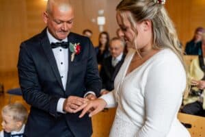 Ein Mann im dunklen Anzug steckt einer Frau während der Hochzeitszeremonie einen Ring an den Finger. Die Frau, strahlend in einem weißen Kleid, ist in dem warm beleuchteten Raum von sitzenden Gästen umgeben. Im Vordergrund beobachtet ein neugieriges Kind den bezaubernden Moment.