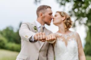 Braut und Bräutigam stehen draußen, küssen sich, strecken eine Hand in die Kamera und zeigen stolz ihre Eheringe. Die Braut in ihrem Spitzenkleid ergänzt den hellen Anzug des Bräutigams perfekt. Dieser romantische Moment fängt die Essenz ihrer sorgfältig geplanten Hochzeit inmitten üppigen Grüns und Bäumen ein.