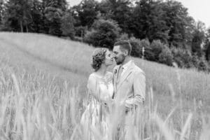 Ein Schwarzweißbild eines Paares in Hochzeitskleidung steht eng beieinander auf einer Wiese. Das Spitzenkleid der Braut und der helle Anzug des Bräutigams schaffen die Kulisse für eine bezaubernde Hochzeit. Bäume säumen den Hintergrund und unterstreichen diese ruhige, romantische Kulisse.