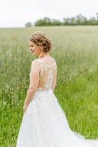 Eine Braut steht auf einer grünen Wiese und trägt ein weißes Spitzenhochzeitskleid mit einem aufwendigen Rückenmuster. Ihr Haar ist in weiche Locken gestylt, während sie über ihre Schulter blickt, umgeben von hohem Gras, das die Schönheit von Hochzeit vor einer verschwommenen Baumreihe in der Ferne verkörpert.