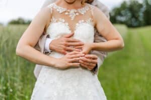 Braut und Bräutigam umarmen sich draußen auf einer üppigen grünen Wiese und feiern ihre Hochzeit. Die Braut trägt ein weißes Kleid mit Spitzenbesatz und sie halten sich an den Händen und zeigen dabei ihre Eheringe. Die Szene fängt einen romantischen Moment mit einem verschwommenen Hintergrund aus Gras und Bäumen ein.