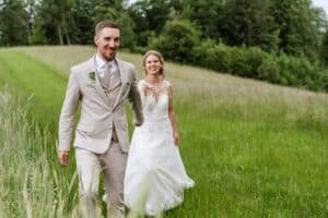 Braut und Bräutigam gehen Hand in Hand über eine Wiese und feiern ihre Hochzeit. Der Bräutigam trägt einen beigen Anzug und die Braut ein weißes Spitzenkleid. Beide lächeln vor einer Kulisse aus grünen Bäumen und einem teilweise bewölkten Himmel, eingefangen in diesem perfekten Moment des Hochzeitsglücks.