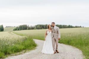 Eine Braut im weißen Kleid und ein Bräutigam im hellbeigen Anzug gehen Arm in Arm einen Kiesweg durch ein üppiges grünes Feld entlang und verkörpern damit die Essenz des Hochzeitsglücks. Sie lächeln sich an, im Hintergrund sind Bäume und ein bewölkter Himmel zu sehen, und fangen die Freude ihres besonderen Tages ein.
