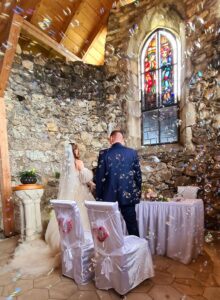 Braut und Bräutigam feiern ihre Hochzeit in einer rustikalen Steinkapelle mit einem atemberaubenden Buntglasfenster. Die Braut strahlt in ihrem weißen Kleid, der Bräutigam sieht in seinem dunklen Anzug schick aus. Die Stühle sind mit weißen Hussen und roten Herzen geschmückt, während Seifenblasen herumschweben und zur fröhlichen Atmosphäre beitragen.