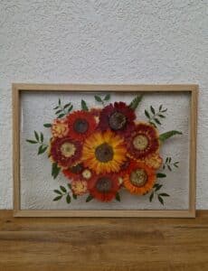 Ein gerahmtes Arrangement aus gepressten, bunten Blumen mit roten, gelben und orangefarbenen Blüten inmitten grüner Blätter verleiht der Hochzeitsplanung eine lebendige Note. Perfekt für die Platzierung vor einer strukturierten weißen Wand auf einer Holzoberfläche, ist es ein ideales Stück für diejenigen, die sich auf ihre Hochzeit vorbereiten.