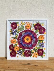 Ein gerahmtes Blumenkunstwerk, perfekt für eine Hochzeit, zeigt eine große zentrale Blume mit bunten Blütenblättern in Lila-, Orange- und Gelbtönen. Dieses lebendige Stück ist von kleineren Blumen und Blättern umgeben und elegant auf einer Holzoberfläche vor einer hell strukturierten Wand präsentiert.