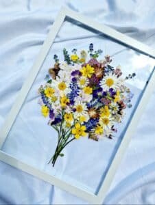 Eine gerahmte Sammlung gepresster Wildblumen, perfekt für die Hochzeitsplanung, zeigt Gänseblümchen und Butterblumen in einem Strauß. Auf einem weichen weißen Stoffhintergrund ruhend, zeigen die Blumen zarte Schattierungen von Gelb, Lila und Weiß – eine charmante Note für jede Hochzeit.