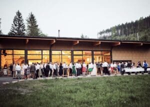 Eine große Gruppe von Menschen versammelt sich vor einem modernen Holzgebäude mit großen Fenstern. Sie sind in formelle Kleidung gekleidet und scheinen an einer Hochzeit teilzunehmen. Die Kulisse besteht aus üppigem grünem Gras und hohen Kiefern unter einem klaren Himmel und bietet eine perfekte Kulisse für diesen besonderen Anlass.