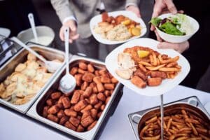 Bei einem Hochzeitsbuffet wird Essen als Teil der durchdachten Hochzeitsplanung serviert. Teller werden mit köstlichen Speisen wie frittierten Kroketten, Pommes Frites und frischem Salat aus großen Metalltabletts auf einem schön gedeckten Tisch gefüllt.